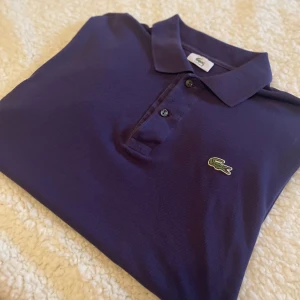 Mörkblå pikétröja från Lacoste - Säljer en klassisk mörkblå pikétröja från Lacoste med den ikoniska gröna krokodilloggan på bröstet. Tröjan har krage, knappar framtill och är tillverkad i bomull. Perfekt för dig som gillar stilren och sportig look.