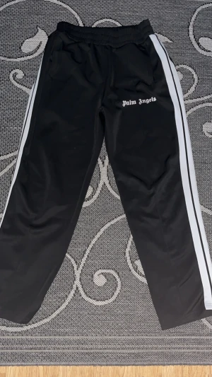 Palm Angels svarta mjukisbyxor med vita ränder - Svarta mjukisbyxor från Palm Angels med vita sidoremsor och logotyp framtill. Byxorna har elastisk midja och raka ben, tillverkade i ett glansigt syntetmaterial som ger en sportig vibe. Perfekta för en chill och trendig streetwear-look. ‼️‼️Är använd några gånger och lite söndrig!!!