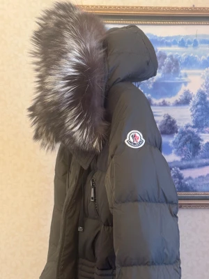 Moncler mörkgrön dunjacka med päls - Snygg mörkgrön dunjacka från Moncler med fluffig päls på huvan. Jackan har långa ärmar, flera dragkedjor framtill och praktiska fickor. Den är quiltad och har en längre passform som ger extra värme. Perfekt för kalla dagar och riktigt stilren look.