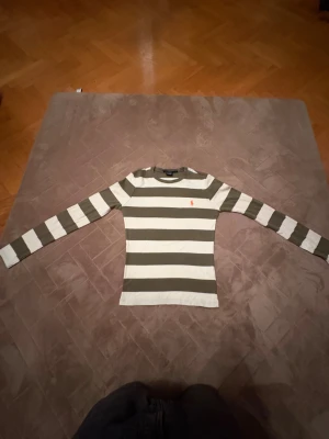 Randig långärmad tröja från Ralph Lauren (Tjej/kvinna) - Snygg långärmad tröja från Ralph Lauren Sport i grönt och vitt med breda ränder. Tröjan har en klassisk rund hals och orange broderad logga på bröstet. Tillverkad i 100% bomull, perfekt för dig som gillar stilrena och sportiga plagg.       (🔥om det syns några fleckar så är det sånt som går att tvätta 🧽 🔥)