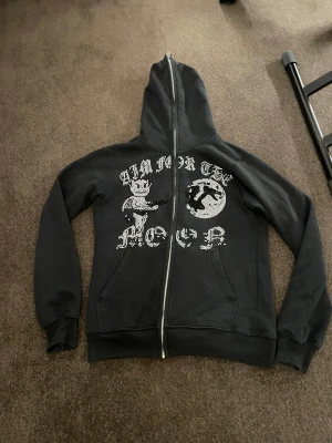 Svart hoodie Aim For The Moon S - Svart hoodie från Aim For The Moon i storlek S med hel dragkedja och huva. Framsidan har ett stort tryck med texten 'Aim For The Moon' och motiv av en tecknad figur och måne i vitt. Tröjan har två fickor fram och ribbade muddar vid ärmslut och nederkant.
