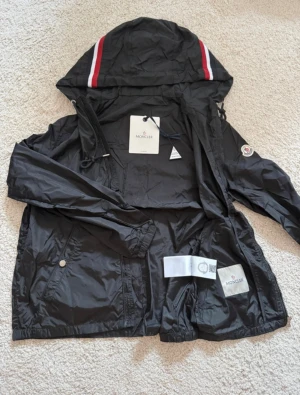 Svart vindjacka från Moncler med huva - Svart vindjacka från Moncler med huva och snygga röda och vita ränder på toppen. Jackan har dragkedja, snörning i huvan och Moncler-logga på ärmen. Materialet är lätt och syntetiskt, perfekt för blåsiga dagar. Fickor med tryckknappar och stilren design.