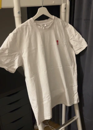 Vit ami Paris t-shirt med rött hjärta - Vit t-shirt från ami Parismed klassisk passform och korta ärmar. På bröstet finns ett broderat rött hjärta med bokstaven A. Tillverkad i mjuk bomull och har även AMI-broderi i nacken. Perfekt basic med snygg detalj.