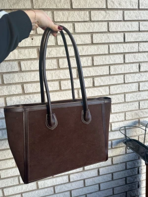 Brun mockaväska - Brun väska i mocka (ej äkta skinn). Praktisk och rymmer en dator mm. Använd ett fåtal gånger. Går att använda antingen med mocka sidan fram eller den glansiga sidan! 