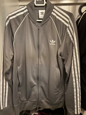 Grå Adidas tröja - Populär Snygg grå tröja från Adidas med klassiska vita ränder längs ärmarna och Adidas-logga på bröstet.  STORLEK L MEN PASSAR M! Byxorna till settet finns i profilen!