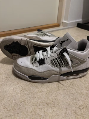 Nike Air Jordan 4 Retro vit/svart - Nike Air Jordan 4 Retro sneakers i vit och svart med klassisk Jumpman-logga på plösen och hälen. Skorna har meshpaneler, snörning och detaljer i mocka och skinn. Snygg platt sula och rund tå, perfekt för dig som gillar streetwear och basketstil.