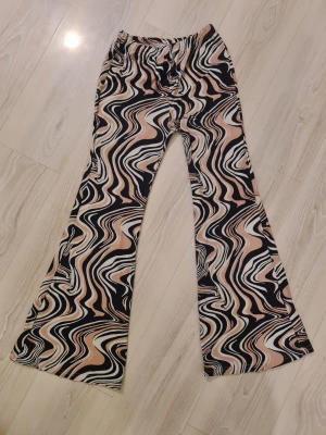 Mönstrade bootcut byxor i rosa, vitt och svart - Säljer ett par riktigt snygga bootcut byxor med swirl-mönster i rosa, svart och vitt. Byxorna har elastisk midja och är tillverkade i ett mjukt, stretchigt material som sitter skönt. Perfekta för dig som vill sticka ut med coola mönster och retrovibbar.