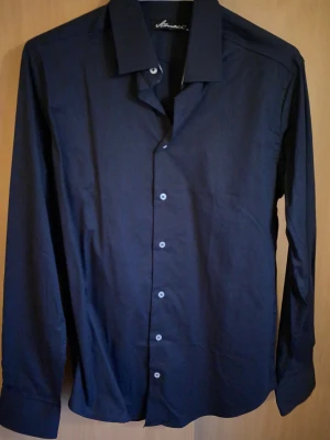 Mörkblå skjorta från Amaci - Snygg mörkblå skjorta från Amaci med klassisk krage och knappar framtill. Skjortan har lång ärm och är tillverkad i mjukt bomullstyg som känns skönt mot huden. Perfekt för dig som gillar stilrena och enkla plagg med en modern touch.