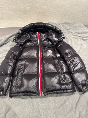Svart Moncler pufferjacka med huva - Säljer en svart pufferjacka från Moncler med blank finish, huva och ikonisk logga på bröstet. Jackan har rödvit dragkedja, två sidofickor med dragkedja och en innerficka med serietidningspatch. Perfekt för kalla dagar och riktigt snygg streetstil.
