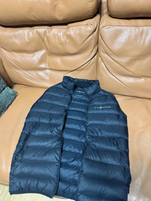 Svart dunväst från Sail Racing - Snygg svart dunväst från Sail Racing med quiltad design och hög krage. Västen har dragkedja framtill och två sidofickor med dragkedja. Perfekt för lager-på-lager och har en stilren logga på bröstet samt en detalj på ryggen. Jag är ca 185cm lång och passar perfekt på mig.