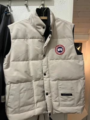 Beige dunväst från Canada Goose - Säljer en beige dunväst från Canada Goose med klassisk logga på bröstet. Västen har hög krage, två stora fickor framtill och svarta tryckknappar. Materialet är slitstark polyester och dunfyllning, perfekt för kalla dagar. Stilsäker och clean design. Kvitto finns inte där av priset. 