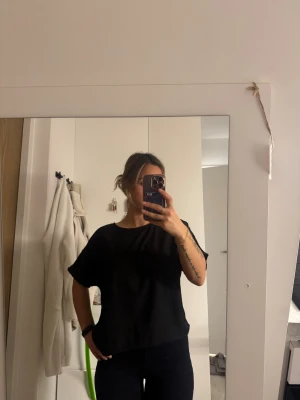 Svart blus med dragkedja bak - En stilren svart blus med rund hals och korta, lösa ärmar. Blusen har en diskret dragkedja i ryggen som ger en cool detalj. Materialet ser ut att vara mjukt och lätt, perfekt för en chill och enkel look.