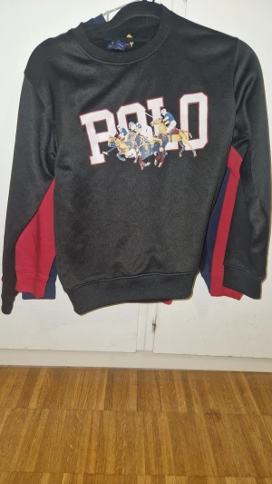 Svart sweatshirt Polo Ralph Lauren - Svart sweatshirt från Polo Ralph Lauren med stort tryck av POLO och polospelare på bröstet. Tröjan har rund hals, långa ärmar och detaljer i rött och blått på sidorna. Tillverkad i mjuk bomullsmix som är skön att bära.