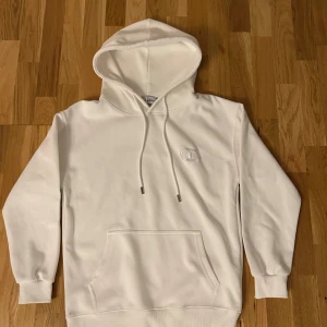 Vit hoodie från Christian Dior - Säljer en clean vit hoodie från Christian Dior med broderad logga på bröstet. Tröjan har huva med snörning, stor magficka och ribbade muddar vid ärmslut och nederkant. Perfekt för dig som gillar stilrena designer och exklusiva märken.