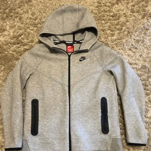 Grå hoodie från Nike XL - Snygg grå hoodie från Nike i storlek XL med svart logga på bröstet. Tröjan har hel dragkedja, två svarta fickor med dragkedja och huva. Materialet är mjukt och känns som bomull och polyester. Perfekt för chill eller träning.