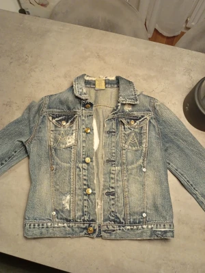 Blå jeansjacka från 7 for all Mankind  - Säljer en klassisk blå jeansjacka från 7 for all Mankind med slitna detaljer och guldiga knappar. Jackan har två bröstfickor med knapp, krage och lång ärm. Materialet är 100% bomull och jackan har en cool vintagekänsla med fransad krage och distressed look.