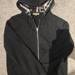 Svart Burberry hoodie med dragkedja - Svart hoodie från Burberry med klassiskt rutigt foder i huvan och snyggt broderad logga på bröstet. Tröjan har snörning i huvan, dragkedja framtill och ribbade muddar. Perfekt för dig som gillar stilrena och exklusiva streetwear-plagg.