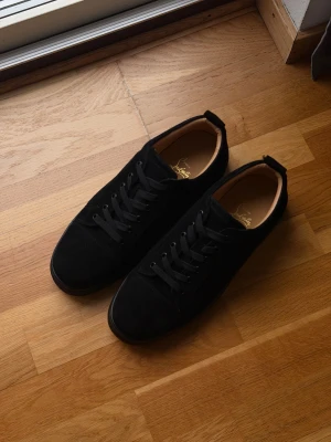 Christian Louboutin svarta utan spikar - Svarta sneakers från Christian Louboutin i mocka med klassisk röd sula och logotyp. Snygg rund tå, svarta snören och lyxig innersula i beige skinn. Kommer med originalpåse, skohorn och box. Perfekt för dig som gillar exklusiv streetstyle. Om någon vill ha fler bilder så är det fritt att fråga! Finns även video ute på vår TikTok! TikTok - ReLuxuria.