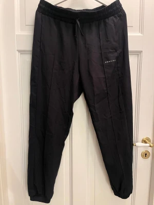 Svarta träningsbyxor från Röhnisch, storlek L - Svarta tunna träningsbyxor från Röhnisch med elastisk midja och snörning. Byxorna har raka ben med resår vid benslut och diskret logga på sidan. Materialet är lätt och syntetiskt, perfekt för kalla dagar. Snygg och enkel design som funkar till många stilar.