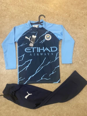 Manchester City långärmad fotbollströja Puma - Snygg Manchester City långärmad fotbollströja från Puma i mörkblått med ljusblå ärmar och blixtmönster. Tröjan har klubbmärke och Etihad Airways-tryck på bröstet. Materialet är lätt och andas, perfekt för träning eller match.