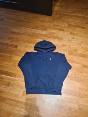 Mörkblå hoodie från Polo Ralph Lauren - Säljer en mörkblå hoodie från Polo Ralph Lauren med klassisk huva och snörning. Tröjan har en broderad logga på bröstet och en stor magficka framtill. Perfekt för dig som gillar stilren och bekväm streetwear.