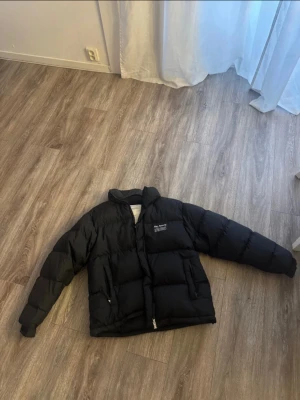 Svart pufferjacka från Axel Arigato - Svart pufferjacka från Axel Arigato med hög krage och två sidofickor med dragkedja. Jackan har en clean look med diskret logga på bröstet och är tillverkad av återvunnen polyester och reprocessed dun. Perfekt för kalla dagar och har en normal passform. Använd 2 gånger 