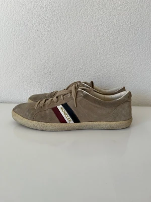 Beige Moncler sneakers i mocka - Snygga beige sneakers från Moncler i mjuk mocka med snörning och rund tå. Skorna är i okej skick då det är tydligt använda och sulan är ganska sliten. Limmet håller på något ställe på att släppa något.