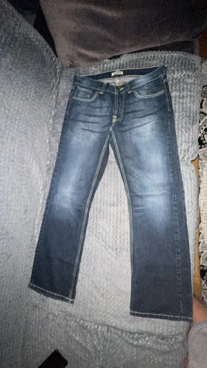 Blå bootcut jeans från Decate W32/L32 - Snygga blå jeans från Decate i klassisk bootcut-modell med kontrastsömmar och fyr fickor. Jeansen har en medelhög Snygga mörkblå jeans i klassisk straight fit-modell. Jeansen har en stilren tvätt med ljusare detaljer framtill som ger ett fräscht och modernt utseende. Bra kvalitet, sköna att ha på sig och passar till allt – t-shirt, hoodie eller skjorta. Perfekta både till vardag och fest. I fint skick utan några skador. och är tillverkade i Italien . Faded effekt framtill och bak ger en cool loo