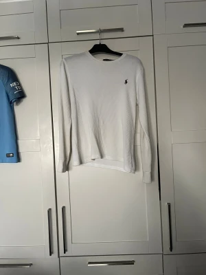 Vit långärmad tröja från Polo Ralph Lauren - Vit långärmad tröja från Polo Ralph Lauren i ribbad bomull. Klassisk rund hals och diskret broderad logga på bröstet. Passar dig som vill ha en clean.