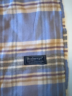Rutig blå halsduk från Burberrys - Snygg rutig halsduk från Burberrys i mjuk lammull. Färgerna går i blått, beige och grått med inslag av lila och vita ränder. Klassisk design med fransar i kanterna, perfekt att slänga över axlarna för en cool look. Made in England.