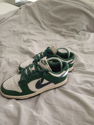Nike Dunk Low gröna och vita sneakers - Snygga Nike Dunk Low sneakers i grönt och vitt med mörkblå swoosh. Skorna har klassisk låg siluett, perforerad tå och gröna snören. Perfekta för dig som gillar streetstyle och vill sticka ut med en fräsch färgkombo.