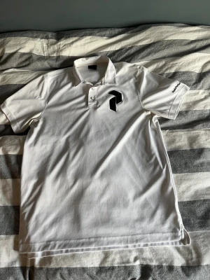 Vit Peak Performance pikétröja - Vit pikétröja från Peak Performance med svart logga på bröstet och broderad text på ärmen. Klassisk krage och knappslå med två vita knappar. Materialet är mjukt och känns sportigt, perfekt för en clean och fräsch look.