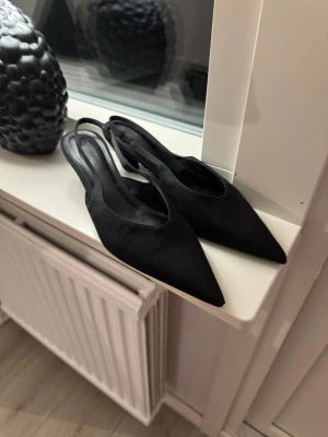 Svarta pumps med spetsig tå från H&M - Snygga svarta pumps från H&M med spetsig tå och låg klack. Skorna har en öppen häl med rem och är tillverkade i ett tygmaterial med diskret struktur. Banden var så när jag köpte de