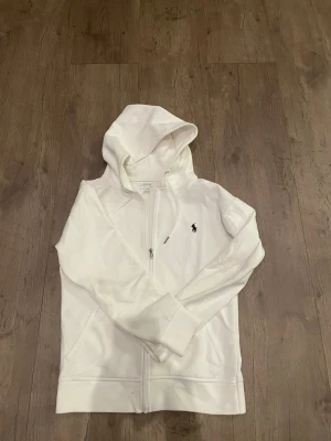 Vit hoodie från Polo Ralph Lauren, storlek S - Vit hoodie från Polo Ralph Lauren med dragkedja och huva. Säljer den då den inte passar längre. Den är riktigt skön och snygg, och väldigt bra skick har inte använts så mycket.Mitt pris är 520kr, Kom med prisförslag!