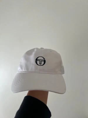 Vit keps från Sergio Tacchini - Vit keps från Sergio Tacchini med svart broderad logga framtill och text baktill. Justerbar rem för perfekt passform. Tillverkad i lätt polyester som andas, perfekt för tennis eller chill dagar. Klassisk böjd skärm och stilren look.