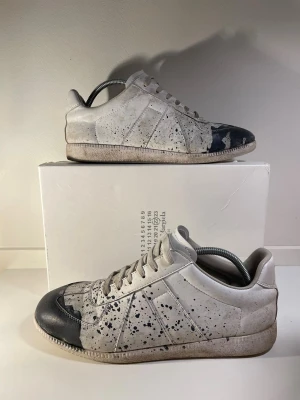 Maison Margiela paint splatter - Unika sneakers från Maison Margiela i vitt skinn med svarta färgstänk och detaljer på tån. Lågt skaft, rund tå och platt sula. Snyggt konstnärligt mönster och klassisk snörning. Perfekt för dig som gillar statement pieces och vill sticka ut.