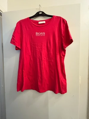 Rosa t-shirt från Hugo Boss - Snygg röd t-shirt från Hugo Boss med klassisk rund hals och vit logotyp på bröstet. T-shirten är kortärmad och tillverkad i mjuk bomull, perfekt för dig som gillar stilrena och trendiga plagg.