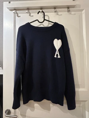 Marinblå stickad tröja från Ami Paris - Säljer en marinblå stickad tröja från Ami Alexandre Mattiussi med vit hjärtlogga och bokstaven A på bröstet. Tröjan har rund hals, ribbade muddar och lång ärm. Materialet är mjuk ull som känns lyxigt och skönt mot huden. Mycket bra skick och sällan använd. Mycket bra skick och sällan använd.