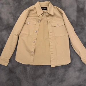 Beige overshirt från Oscar Jacobson - Stilren beige overshirt från Oscar Jacobson i bomull. Skjortan har klassisk krage, två bröstfickor med lock och knappar samt långa ärmar. Perfekt lager-på-lager-plagg med snygg passform och diskreta detaljer.