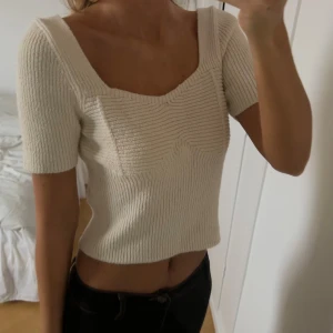 Beige croppad stickad topp - Säljer en beige croppad stickad topp med korta ärmar och fyrkantig halsringning. Toppen har ribbad struktur och markerade sömmar vid bysten som ger en snygg form. 