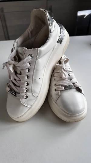 Vita sneakers från Steve Madden - Snygga vita sneakers från Steve Madden med silvriga detaljer på hälen och metallplatta med logga på snörningen. Skorna har rund tå, platt sula och är tillverkade i skinn med textilfoder. Perfekta för en clean och trendig look.