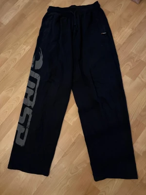 Svarta Gasp mjukisbyxor med tryck - Svarta mjukisbyxor från gasp med bred passform och stort grått tryck längs ena benet. Byxorna har snörning i midjan och är gjorda i mjukt bomullsmaterial, perfekta för chill eller sport. Snyggt street-inspirerat utseende.