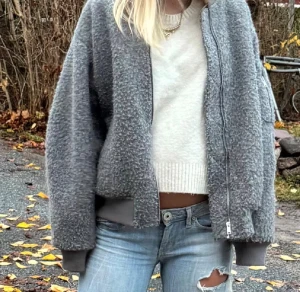 Grå teddy bomberjacka i storlek L - Säljer en grå bomberjacka med ribbade muddar och krage. Jackan har en grov, fluffig yta och stängs med dragkedja framtill. Modellen är loose fit och har långärmade ärmar. 