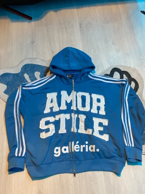 Blå hoodie Stile Amor  - Blå hoodie med vita tryck och broderade bokstäver framtill och baktill. Klassiska vita ränder längs ärmarna, huva och dragkedja hela vägen. Oversized vibe och riktigt streetwear-stil. Materialet är mjuk bomullsmix. Perfekt för dig som gillar statement-plagg. Äkta har kvitto för den som vill se