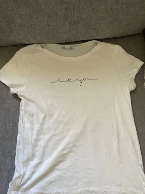 Vit t-shirt från Amisu med tryck - Säljer en vit t-shirt från Amisu i storlek S med ett stilrent svart tryck på bröstet där det står 'i love you' med ett hjärta. T-shirten har rund hals och korta ärmar. Tröjan har en liten fläck, kanske går att få bort. 