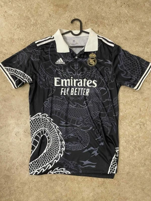 Real Madrid svart draktröja Adidas - Säljer en svart Real Madrid fotbollströja från Adidas med fet drakdesign över hela tröjan. Vit krage och vita detaljer på ärmarna, samt klubbmärke och sponsortryck på bröstet. Materialet är lätt och andas, perfekt för match eller träning.
