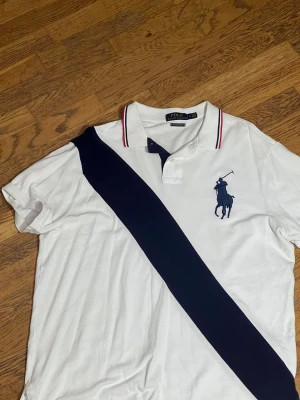 Vit Polo Ralph Lauren piké slim fit - Vit pikétröja från Polo Ralph Lauren med marinblå diagonal rand och stor broderad polospelare på bröstet. Slim fit-modell med klassisk krage och röda/blå detaljer på kragen. Tillverkad i mjuk bomull, perfekt för en clean och sportig look. Den kan vara lite stor i storleken ligger mellan m/L men mycket mer åt M hållet. 
