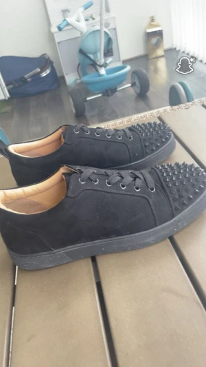 Christian Louboutin svarta sneakers med nitar - Svarta sneakers från Christian Louboutin i mocka med coola svarta nitar på tån och klassisk röd sula med logga. Skorna har snörning och rund tå, samt beige innersida i skinn. Perfekt för dig som gillar edgy och lyxig stil.