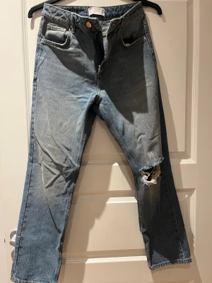 Blå jeans med hål från ASOS Design - Säljer ett par blå jeans från ASOS Design med klassisk femficksmodell och ett snyggt hål på ena knät. Jeansen har raka ben och normal passform, tillverkade i bomull med dragkedja och knapp framtill. Perfekta för en avslappnad och trendig look.