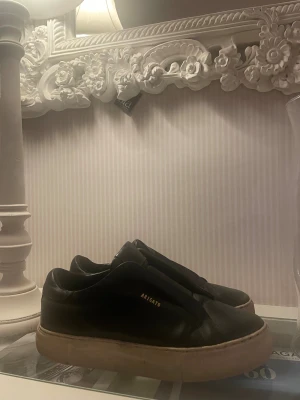 Svarta sneakers från Axel Arigato - Svarta sneakers från Axel Arigato med stilren design och guldfärgad logga på sidan. Skorna har en vit platt sula och är tillverkade i skinn. Rund tå och elastisk panel istället för klassisk snörning ger en clean look.